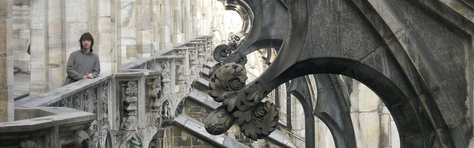 A student stands on a balcony in a stone building, which has stone arches, balconies, and pillars with intricately carved details that repeat about every six feet. 